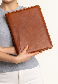 Carpeta de cuero marrón con una textura suave, con un borde curvado y costuras en contraste. Sostenida en manos frente a un fondo claro.