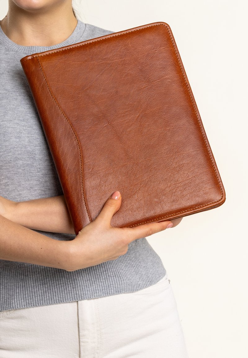 Carpeta de cuero marrón con una textura suave, con un borde curvado y costuras en contraste. Sostenida en manos frente a un fondo claro.