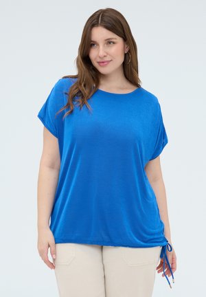 Frau mit langen braunen Haaren, die ein knallblaues, kurzärmeliges Oberteil mit seitlicher Schleife und beige Hosen trägt, steht vor einem schlichten Hintergrund.