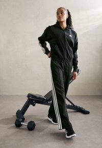 Svart atletisk träningsoverall med vita ränder, bestående av en zip-up huvtröja och tapered byxor, bärs med svart och vita sneakers bredvid gymutrustning.