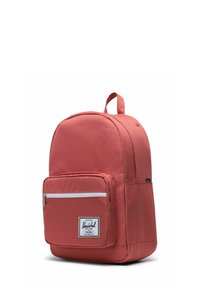Sac à dos de couleur corail avec une poche avant zippée, un tissu lisse, une forme arrondie et une poignée supérieure. Présente un patch logo sur le devant.