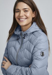 Veste matelassée bleu clair avec capuche, dotée d'une fermeture éclair et de cordons de serrage noirs. Logo brodé sur la manche gauche. Texture douce.