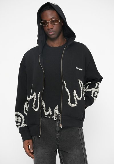 Άνδρας φορώντας μαύρη κουκούλα με λευκά σχέδια φλόγας, μαύρο t-shirt, μαύρα τζιν, ζώνη με καρφιά και ορθογώνια γυαλιά ηλίου, με τα χέρια στις τσέπες.