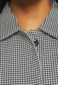 Tessuto a motivo pied de poule in bianco e nero con colletto appuntito e un unico bottone nero al collo. Texture liscia, design aderente.