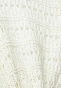 Manta de ganchillo en blanco, con un tejido abierto texturizado que presenta patrones intrincados y tamaños de punto variados, cayendo de manera suelta.