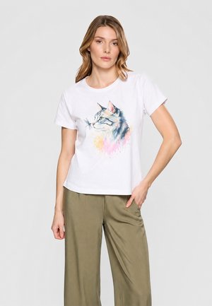 Mujer con camiseta blanca con estampado de gato y libélula estilo acuarela, combinada con pantalones sueltos verde oliva, de pie frente a un fondo liso.