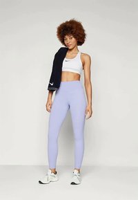 Bela športna modrček, lilaste legice in bele superge; nosi temno brisačo čez ramo. Gladek material in prilegajoč dizajn.