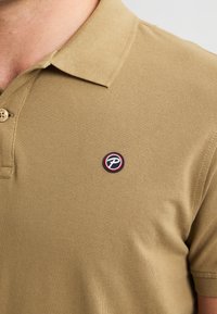 Polo shirt tan realizzato in tessuto testurizzato. Caratterizzato da un colletto classico e una toppa con logo sulla parte superiore sinistra, racchiusa da un design circolare.