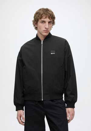 Man met lichtbruin krullend haar, die een zwart jack met rits draagt met geribde boorden en een klein vierkant logo op de borst.