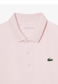 Koszulka polo w jasnoróżowym kolorze, z krótkimi rękawami i płaskim kołnierzykiem. Posiada dwie guziki przy wykroju i haftowane zielone logo krokodyla na lewej piersi.