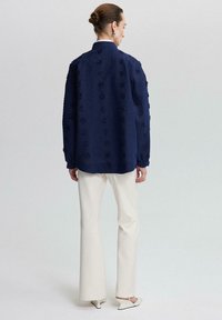Navy blue textured shirt with floral pattern, long sleeves, button closure, paired with white flared pants and white slip-on shoes.