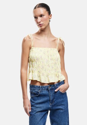 Mujer que lleva un top sin mangas amarillo con estampado floral y tirantes ajustables, y vaqueros azules de tiro alto, posando con una mano en el bolsillo.