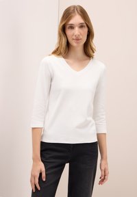 Weißes, tailliertes Langarm-T-Shirt mit V-Ausschnitt und weicher Textur, kombiniert mit schwarzen Jeans. Einfaches Design ohne Muster oder Akzente.