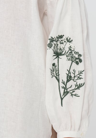 Linned skjorte i myk hvit med lange ermer. Har grønn blomsterbroderi på den ene ermen og en knappmansjett. Teksturert, lett stoff.