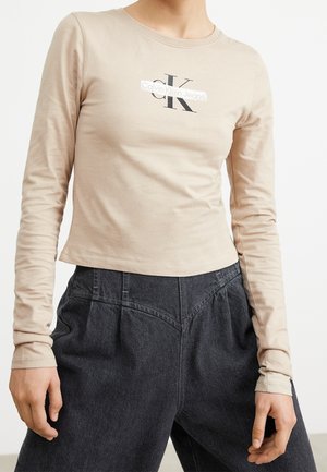 Osoba ubrana w beżową bluzkę Calvin Klein Jeans z długim rękawem, włożoną w wysokie, luźne czarne spodnie dżinsowe, na białym tle.