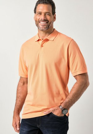 Polo de manga corta en algodón naranja claro, con cuello clásico, tapeta de tres botones y ajuste relajado. Combinado con vaqueros de mezclilla.