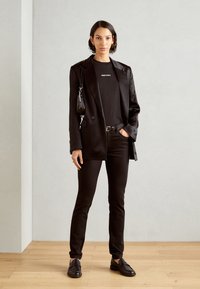 Zwarte blazer met een gladde textuur over een zwart T-shirt, gecombineerd met strakke zwarte broek en zwarte loafers. Kleine handtas met schouderband.