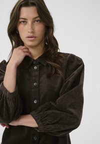Brown corduroy shirt jacket with a collared neckline, button closure, and voluminous puff sleeves. Textured fabric and black buttons.