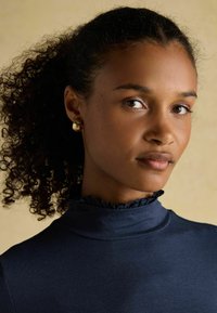 Korts, riides kuldsete rõngastega, tumesinise kõrge kraega rüüga naine, kelle lokkis juuksed on tagasi tõmmatud, neutraalse beeži tausta ees.