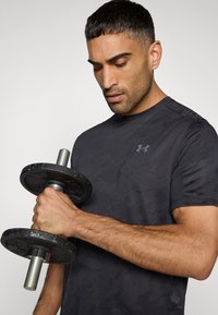 Μαύρο αθλητικό πουκάμισο με υφασμένο σχέδιο, με το λογότυπο της Under Armour. Το άτομο κρατάει ένα μαύρο αλτήρα με ασημένιες λεπτομέρειες.