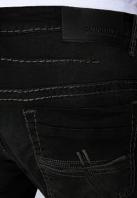 Los jeans de mezclilla negra presentan un acabado texturizado, costuras en gris contrastante y una etiqueta de cuero en la cintura. La parte trasera incluye detalles en los bolsillos.