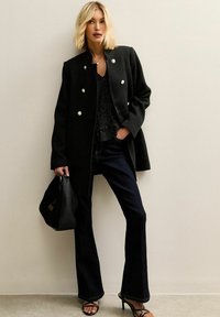 Zwarte wollen jas met zilveren knopen, gecombineerd met een zwart met witte stippen top, donkerblauwe flared jeans en zwarte sandalen met hakken. Zwarte handtas inbegrepen.