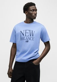 Άνδρας με φανέλα New Balance ανοιχτού μπλε χρώματος, όρθιος με το ένα χέρι στην τσέπη μαύρου παντελονιού, κοιτάζοντας προς τα δεξιά του πάνω σε απλό φόντο.