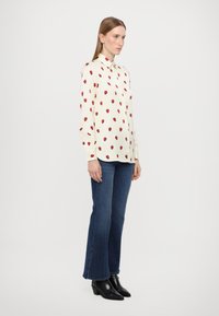 Weißes Hemd mit rotem Erdbeer-Muster, lange Ärmel und Kragen, kombiniert mit ausgestellten blauen Jeans und schwarzen Stiefeletten.