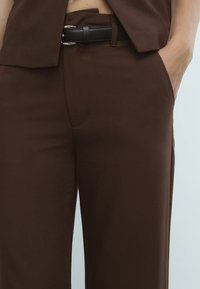 Hnědé tailored kalhoty vyrobené z hladké tkaniny, s přiléhavým střihem, skrytými kapsami a černým opaskem se stříbrnými detaily.