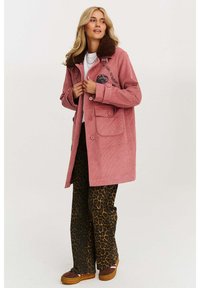 Ružový kordový kabát s hnedým kožúškom a vyšitým logom, s veľkými vreckami. V kombinácii s nohavicami s leopardím vzorom a hnedými topánkami.