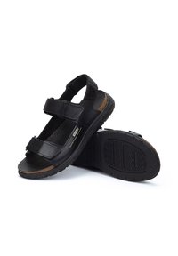 Sandalias de cuero negro con dos correas ajustables, plantilla texturizada, suela de goma y un acento marrón en el lateral; diseñadas para la comodidad y la durabilidad.