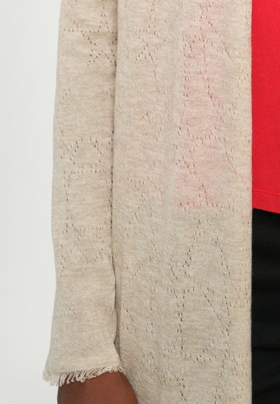 Beige cardigan with a knitted pattern, featuring a loose fit, frayed cuffs, and a soft texture, layered over a red top.
