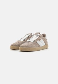Une paire de baskets en daim beige avec des lacets blancs, des rayures blanches sur les côtés et des semelles en caoutchouc marron clair sur un fond blanc.