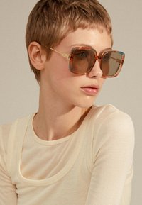 Occhiali da sole oversize in acrilico con una sfumatura ambrata e una montatura trasparente marrone. Presentano dettagli in metallo dorato e bordi squadrati.