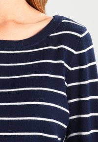 Námořnická modrá pletená svetr s bílými horizontálními pruhy; má široký výstřih a žebrovanou texturu na rukávech a lemu.