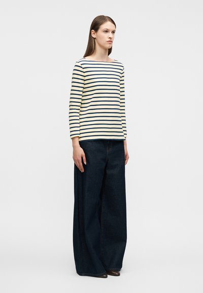 Γυναίκα που φοράει μπλούζα με άσπρες και ναυτικές ρίγες μακριάς μανίκια και άνετο σκούρο μπλε τζιν παντελόνι με φαρδιά μπατζάκια, στέκεται μπροστά σε λευκό φόντο.