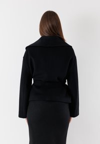 Giacca nera in lana con un ampio colletto, maniche lunghe, vita alta con cintura e silhouette strutturata. Indossata sopra una gonna nera aderente.