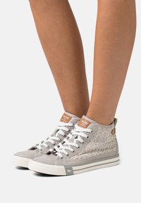 Světle šedé vysoké tenisky s texturou šněrování, bílé tkaničky a hnědé kožené akcenty. Gumová podrážka s logem na boku.