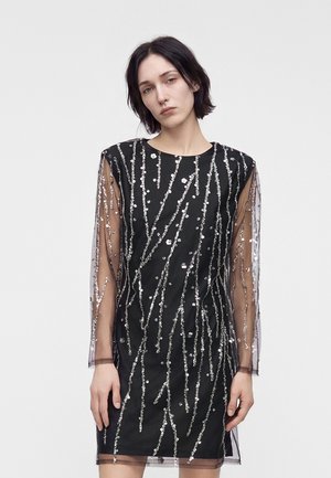 Jeune femme aux cheveux courts et foncés porte une robe noire ornée de sequins argentés et de manches longues transparentes sur un fond clair uni.