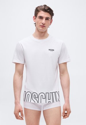 Giovane uomo che indossa una t-shirt bianca Moschino Active con logo nero e slip bianchi abbinati, in piedi davanti a uno sfondo chiaro semplice.