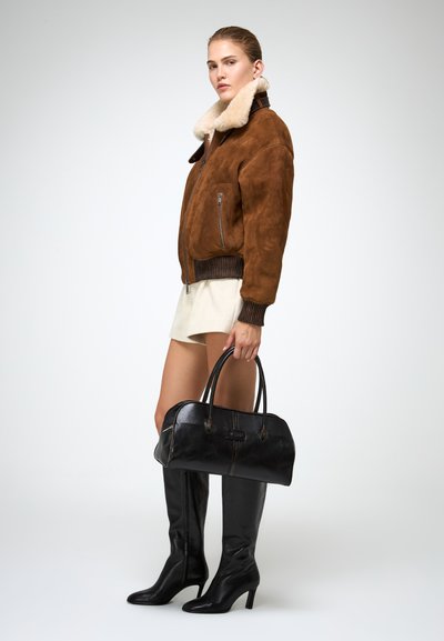Giacca bomber in suede marrone con colletto in pelliccia, pantaloni corti color crema, stivali neri in pelle al ginocchio e una borsa nera con manici arrotondati.