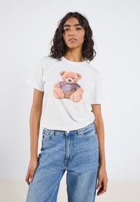 Hvit bomulls t-skjorte med et mykt teddybjørn-trykk med lilla skjorte. Modellen har på seg lyseblå denimjeans. Enkelt design og avslappet passform.