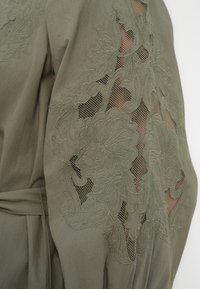 Ενδυμα σε ελιά πράσινο χρώμα με περίτεχνη κεντητή λουλουδένια διακόσμηση και λεπτομέρειες από δίχτυ στο μανίκι, με ζώνη στη μέση και μαλακή υφή υφάσματος.