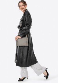 Cappotto di pelle nera, borsa a tracolla grigia texturizzata, pantaloni grigi chiari e tacchi neri con dettaglio a fiocco. Il modello sta in profilo, sorridendo.
