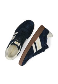 Granatowe zamszowe sneakersy o niskim profilu, z kremowymi paskami i logo, brązową gumową podeszwą oraz teksturowaną cholewką.