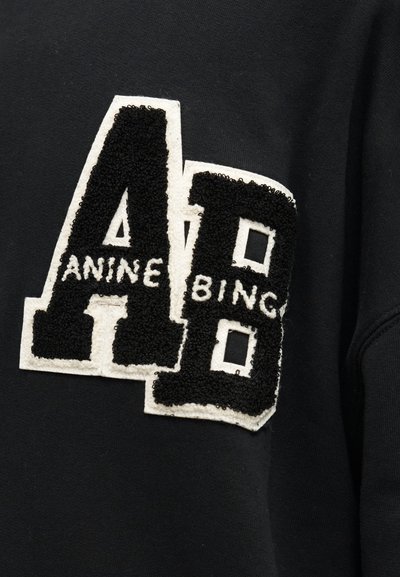 Fekete pulóver texturált fekete-fehér betűpatch-csel, ahol az "A" és "B" betűk átfedésben vannak, a mellrészen részlegesen olvasható "ANINE BING" felirattal.