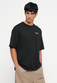 Crna pamučna majica s kružnim izrezom, kratkim rukavima i malim bijelim Calvin Klein logotipom na lijevoj strani prsnog koša. Ravan rub.