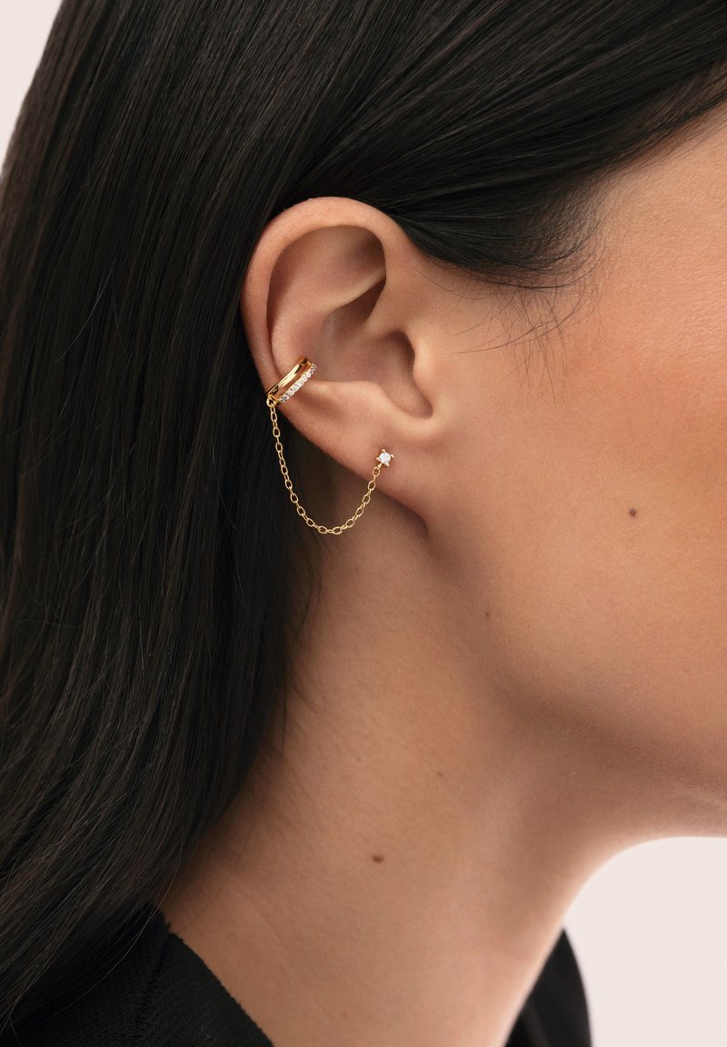 Gros plan d'une oreille portant deux boucles d'oreilles en or reliées par une délicate chaîne en or, une puce et un bracelet ornés de petits cristaux.