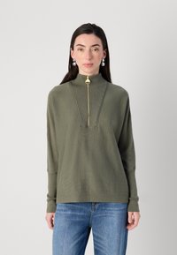 Model trägt einen grünen, halb mit Reißverschluss versehenen Pullover mit einem hohen Kragen, kombiniert mit blauen Jeans, stehend vor einem neutralen Hintergrund.