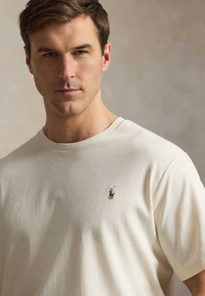 Rövid barna hajú férfi, aki egyszínű bézs pólót visel, melyen a mellrészen kis hímzett pólló játékos logó található.